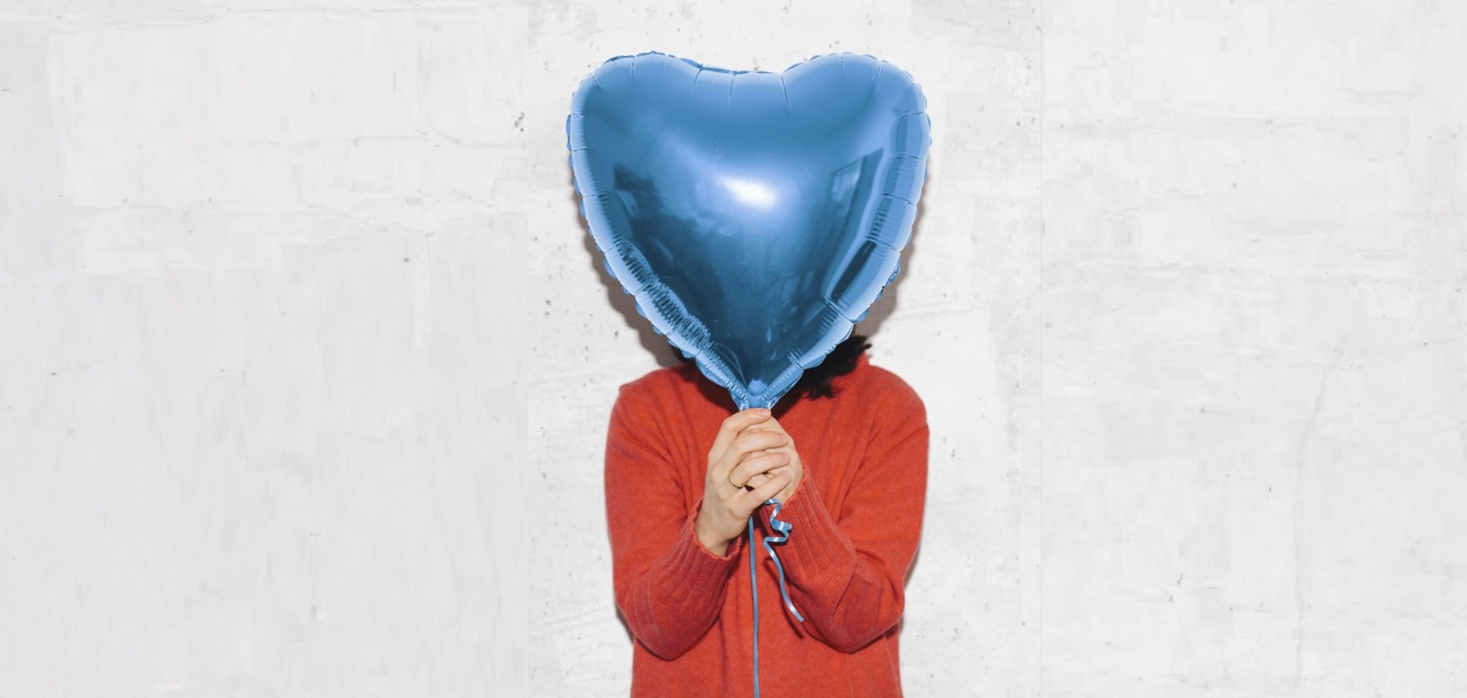 NetCologne Engagement Person in rotem Pullover hält einen großen blauen Herzballon vor einer hellen Betonwand.