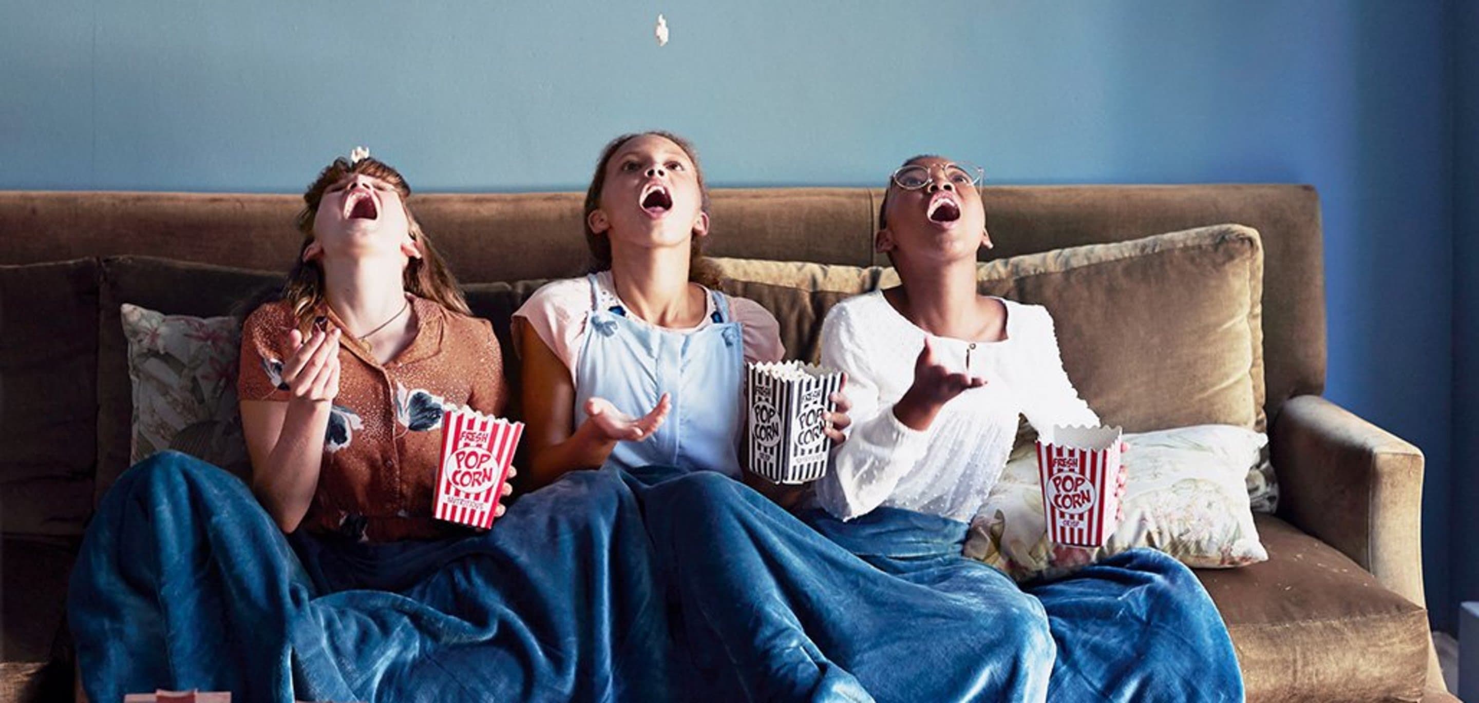 Drei Mädchen auf einer Couch werfen Popcorn in die Luft, und versuchen es mit dem Mund aufzufangen