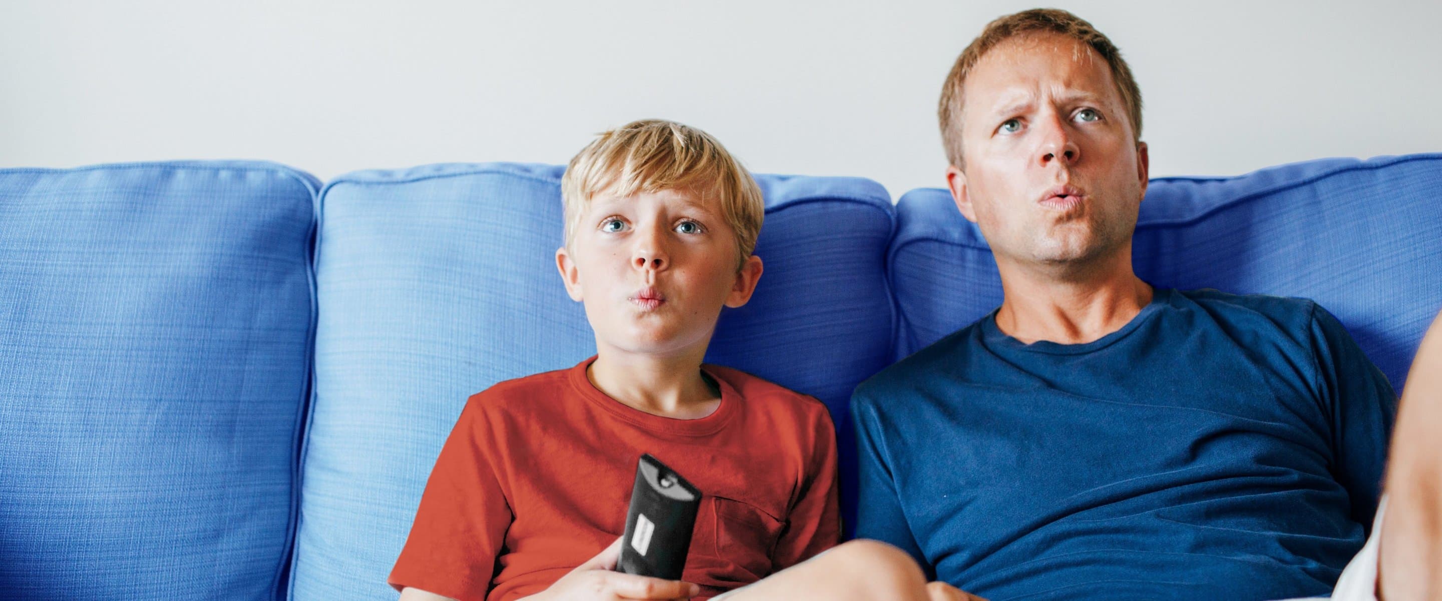 Vater und Sohn, der Fernbedienung in der Hand hält, sitzen auf einer Couch und sehen erstaunt aus