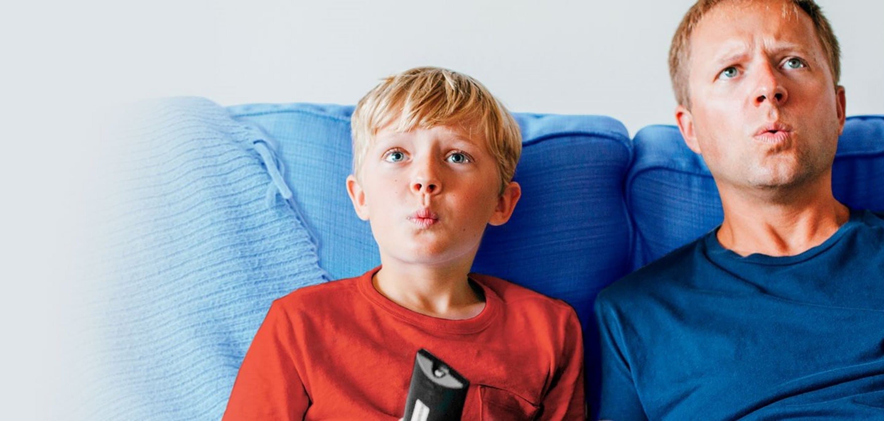 Vater und Sohn, der Fernbedienung in der Hand hält, sitzen auf einer Couch und sehen erstaunt aus