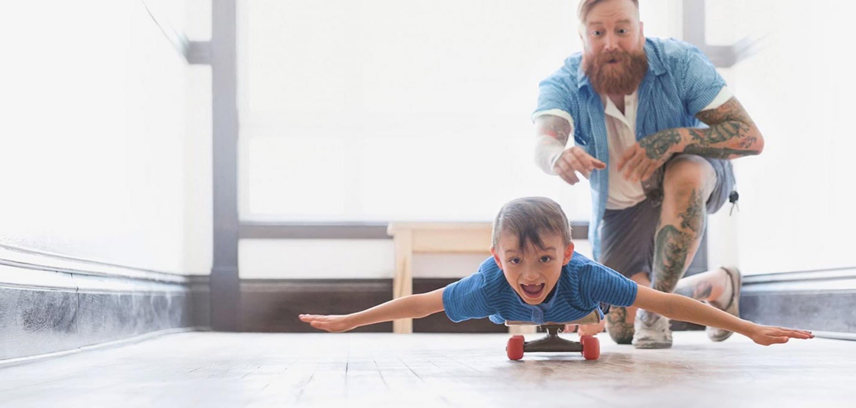 Vater schubst Sohn an, der mit ausgebreiteten Armen auf einem Skateboard liegt.
