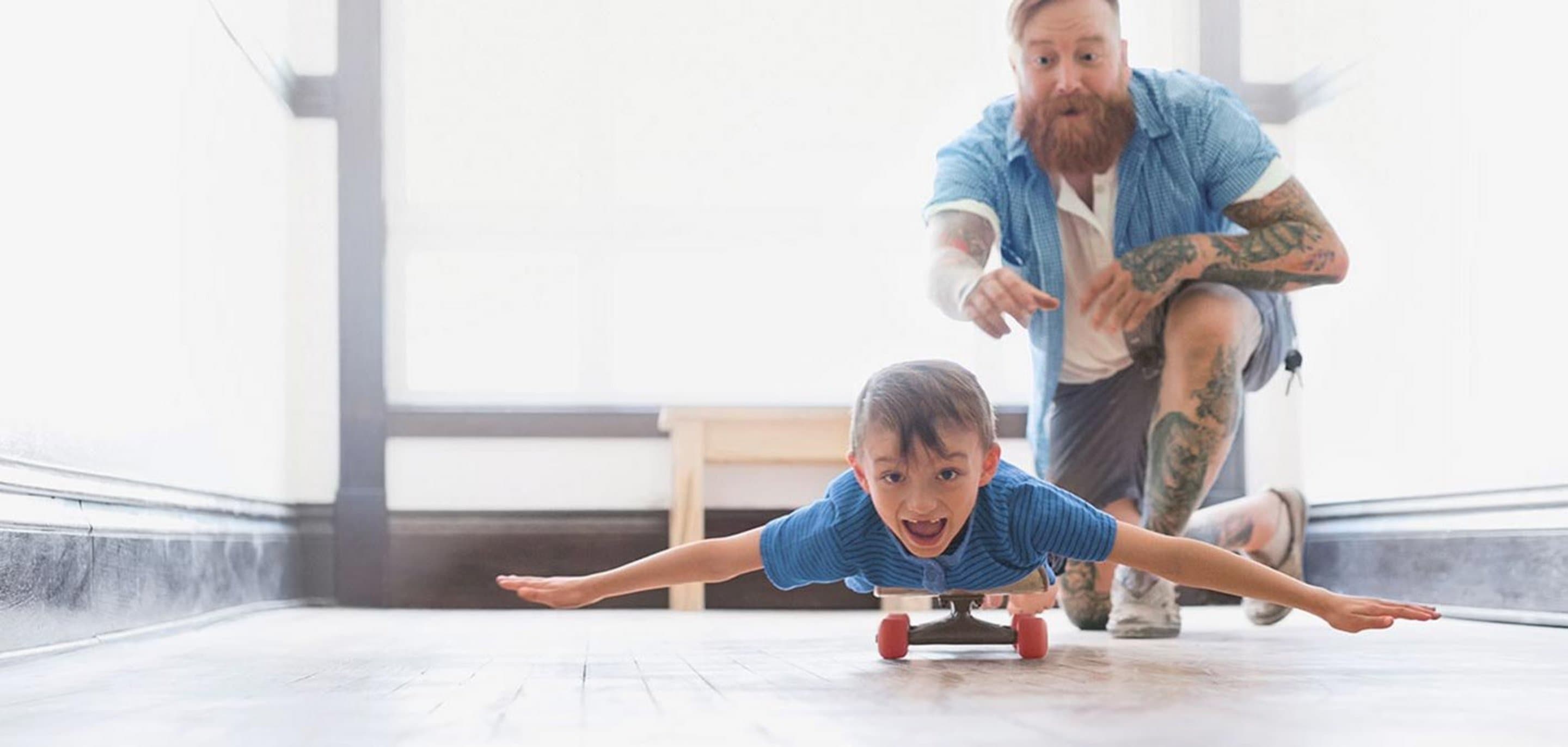 Vater schubst Sohn an, der mit ausgebreiteten Armen auf einem Skateboard liegt.