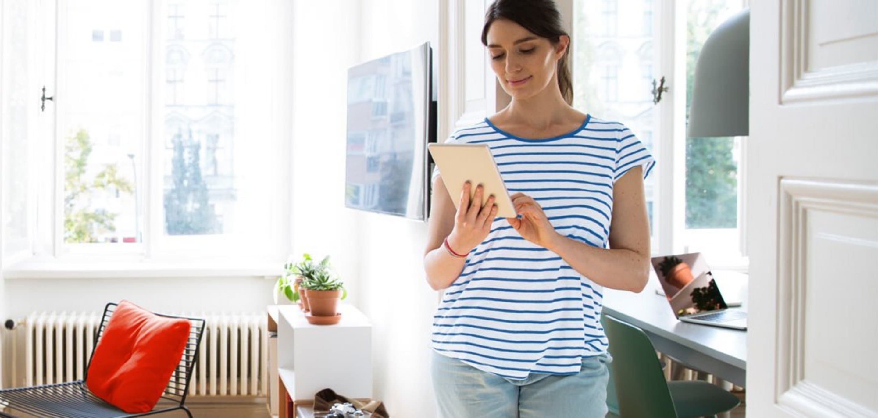 Person steht in einem hellen, modernen Wohnzimmer und hält ein Tablet in der Hand; Szene symbolisiert Zufriedenheit aufgrund einer guten Internetverbindung und starkem WLAN-Signal.