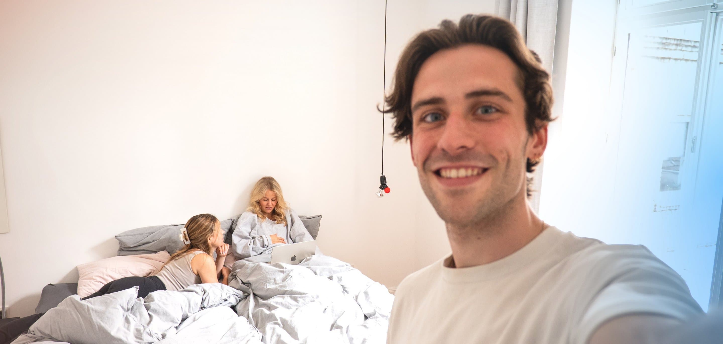 In einem Schlafzimmer liegen zwei Personen auf einem Bett und schauen gemeinsam auf ein Laptop, eine dritte Person steht im Vordergrund und macht ein Selfie.