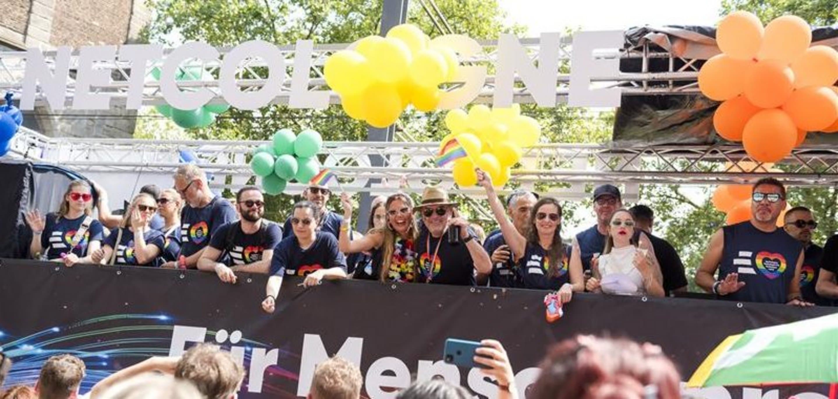 NetCologne-Paradewagen mit bunten Ballons und Menschen in Shirts mit Regenbogenherz, die fröhlich winken. Teil einer LGBTQ+ Pride-Parade.