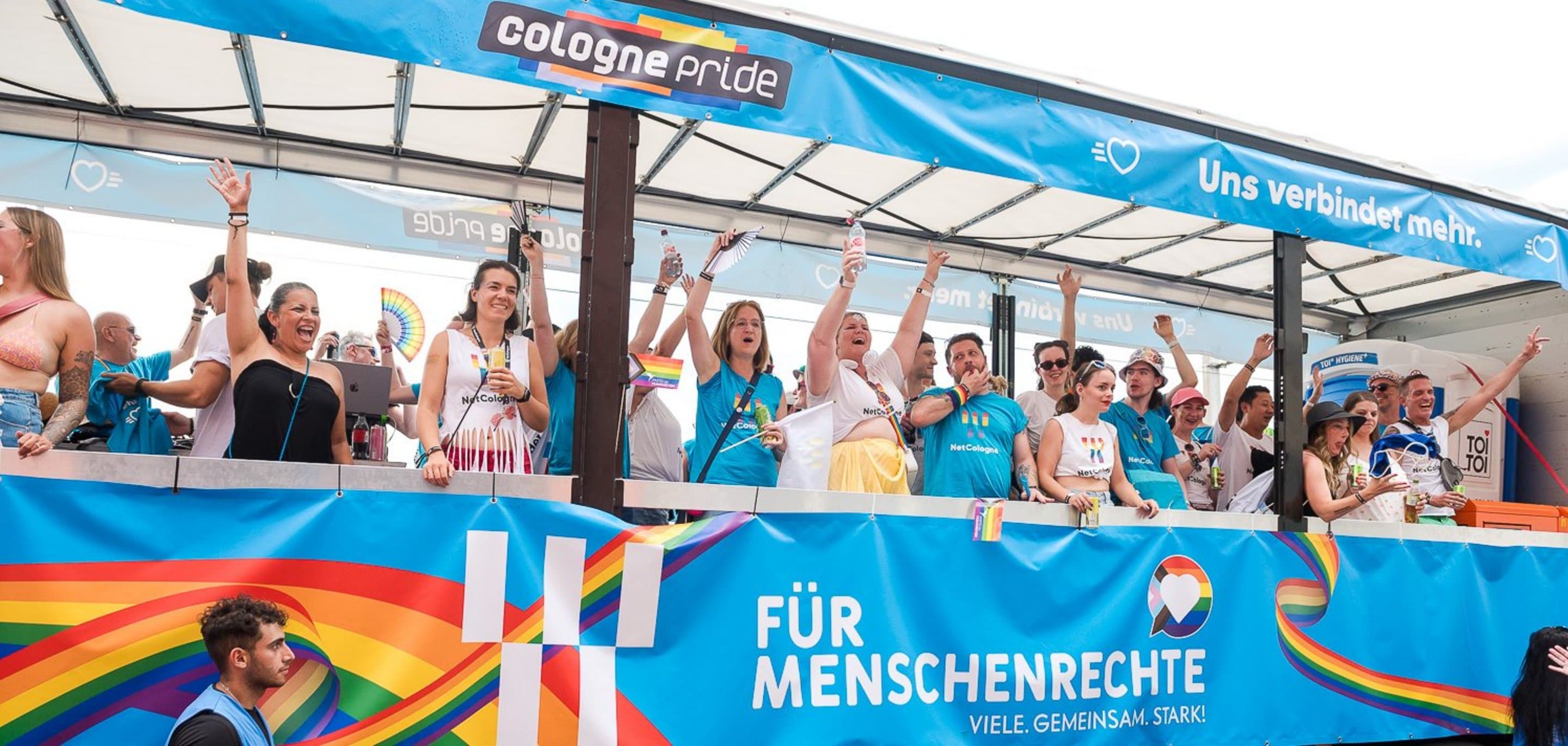 Paradewagen von NetCologne mit Regenbogenfarben und dem Slogan ‚Für Menschenrechte – Viele. Gemeinsam. Stark!‘, begleitet von fröhlich winkenden Menschen.
