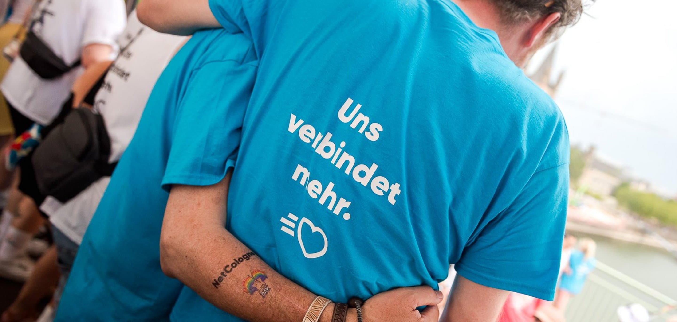 Zwei Personen von hinten in blauen T-Shirts, eine mit Regenbogen-Tattoo "NetCologne". Auf dem Rücken steht: "Uns verbindet mehr."