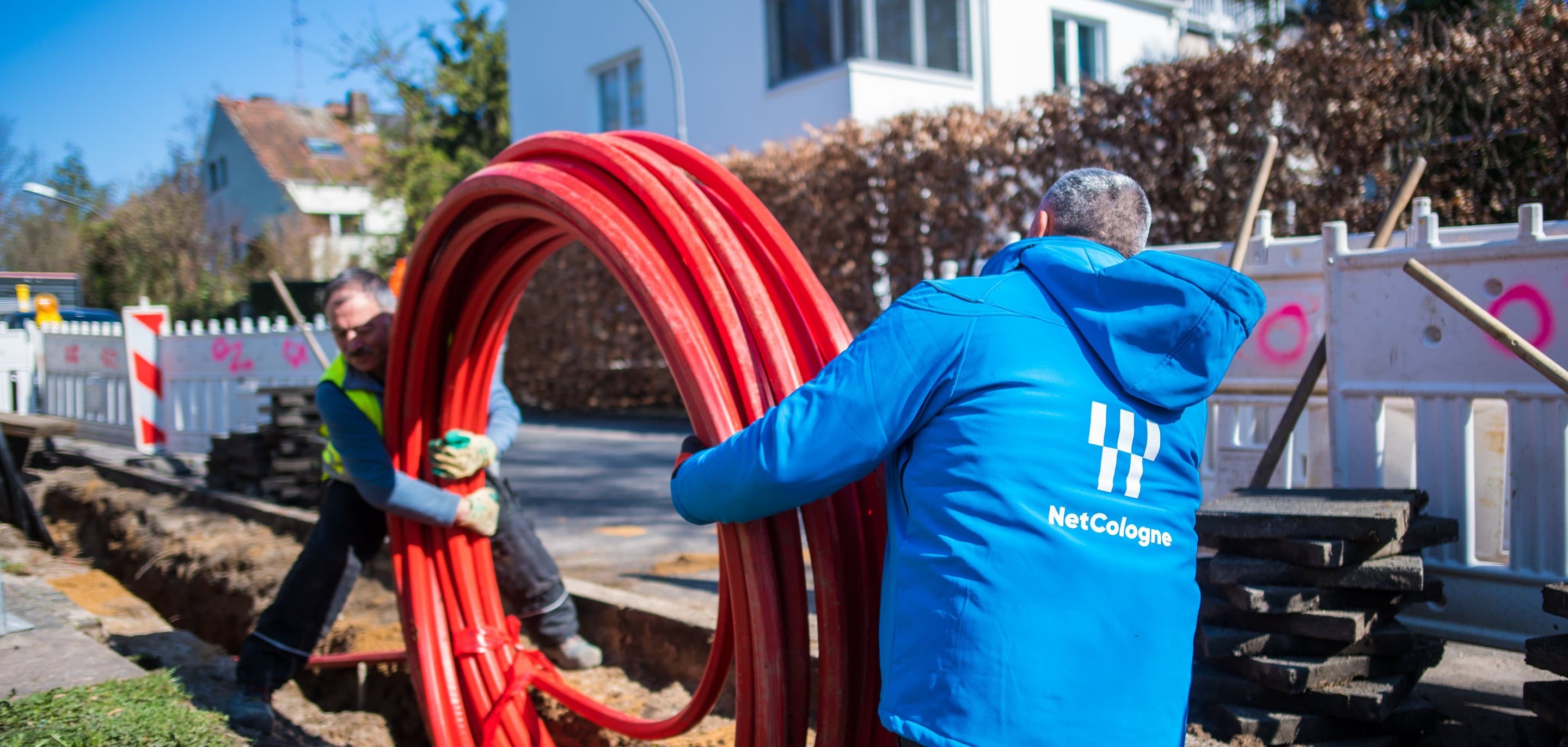 NetCologne-Mitarbeiter in blauer Jacke halten aufgerollte rote Glasfaserkabel-Leerrohre an einer Straßenbaustelle.
