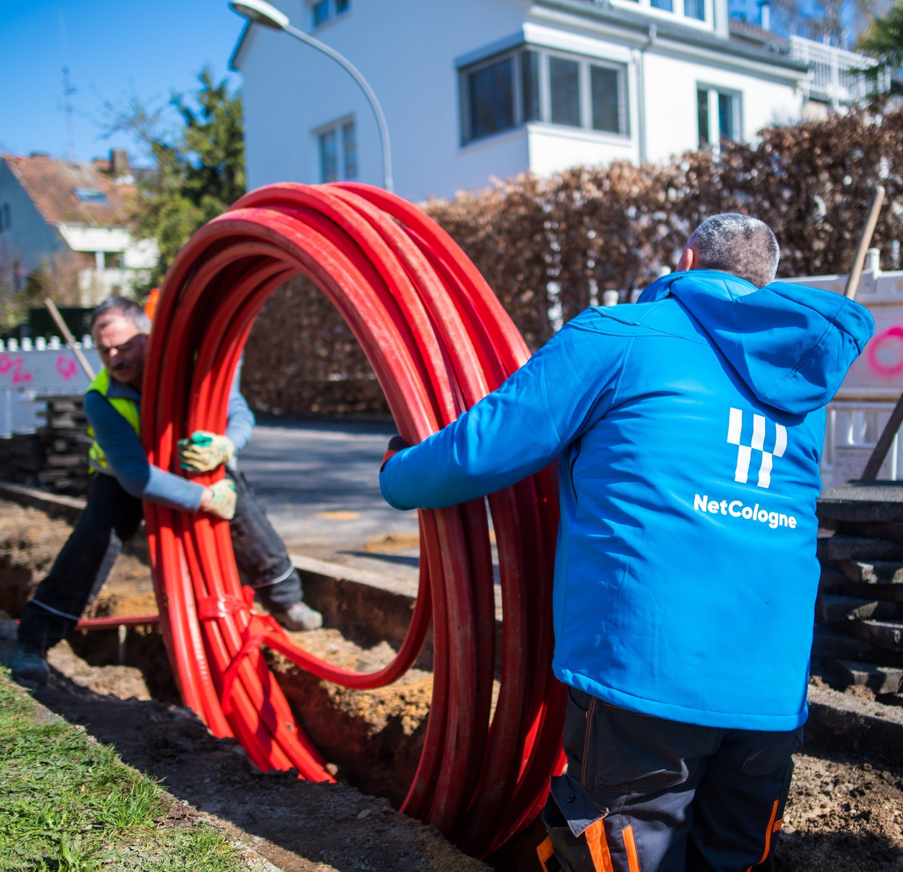 NetCologne-Mitarbeiter in blauer Jacke halten aufgerollte rote Glasfaserkabel-Leerrohre an einer Straßenbaustelle.