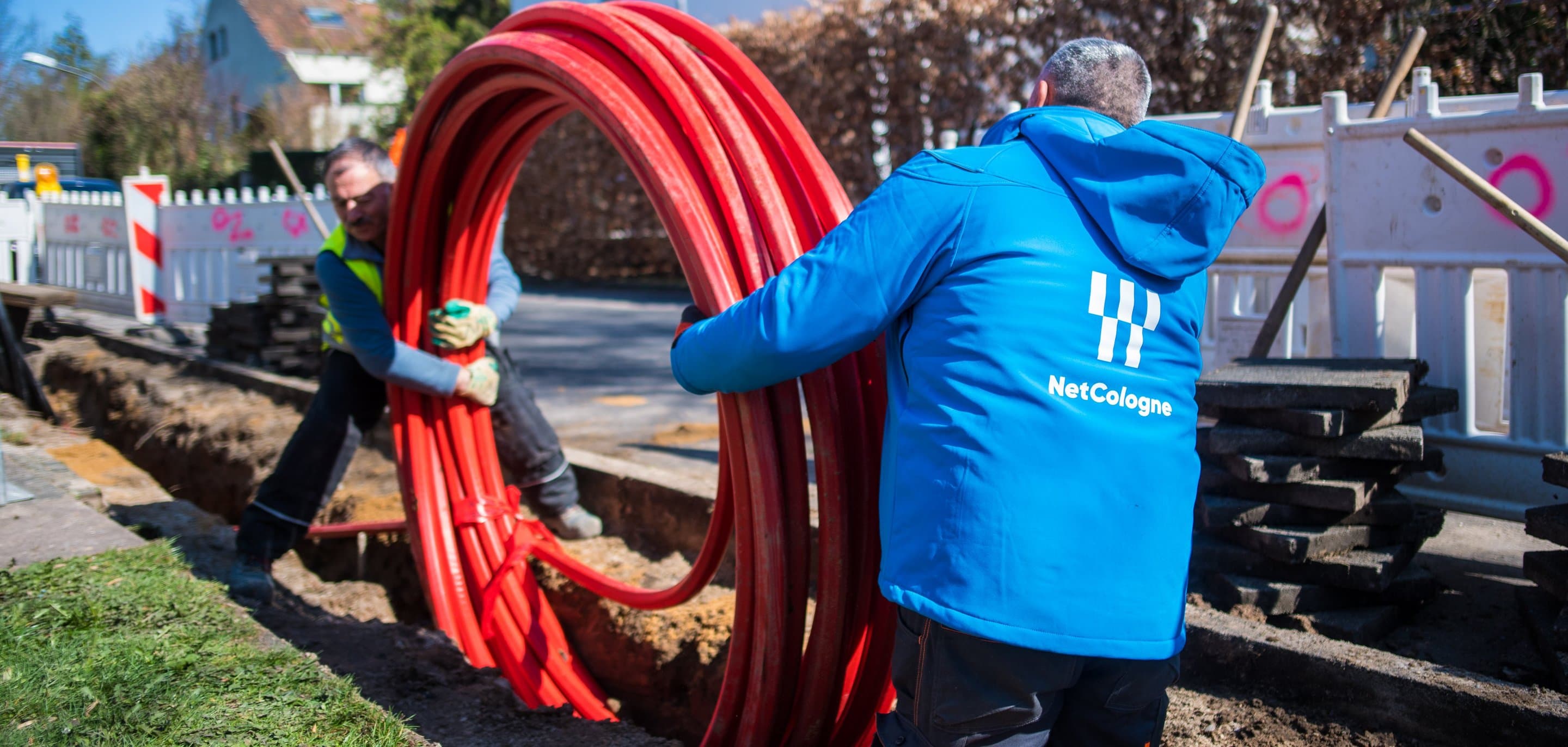 NetCologne-Mitarbeiter in blauer Jacke halten aufgerollte rote Glasfaserkabel-Leerrohre an einer Straßenbaustelle.