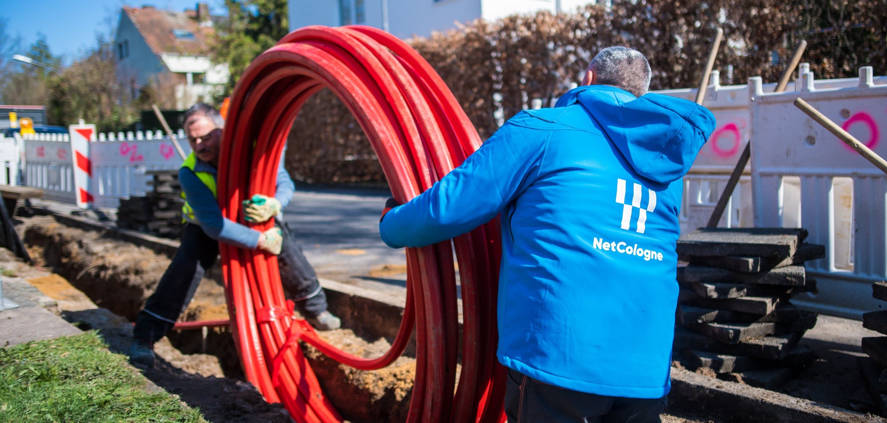 NetCologne-Mitarbeiter in blauer Jacke halten aufgerollte rote Glasfaserkabel-Leerrohre an einer Straßenbaustelle.