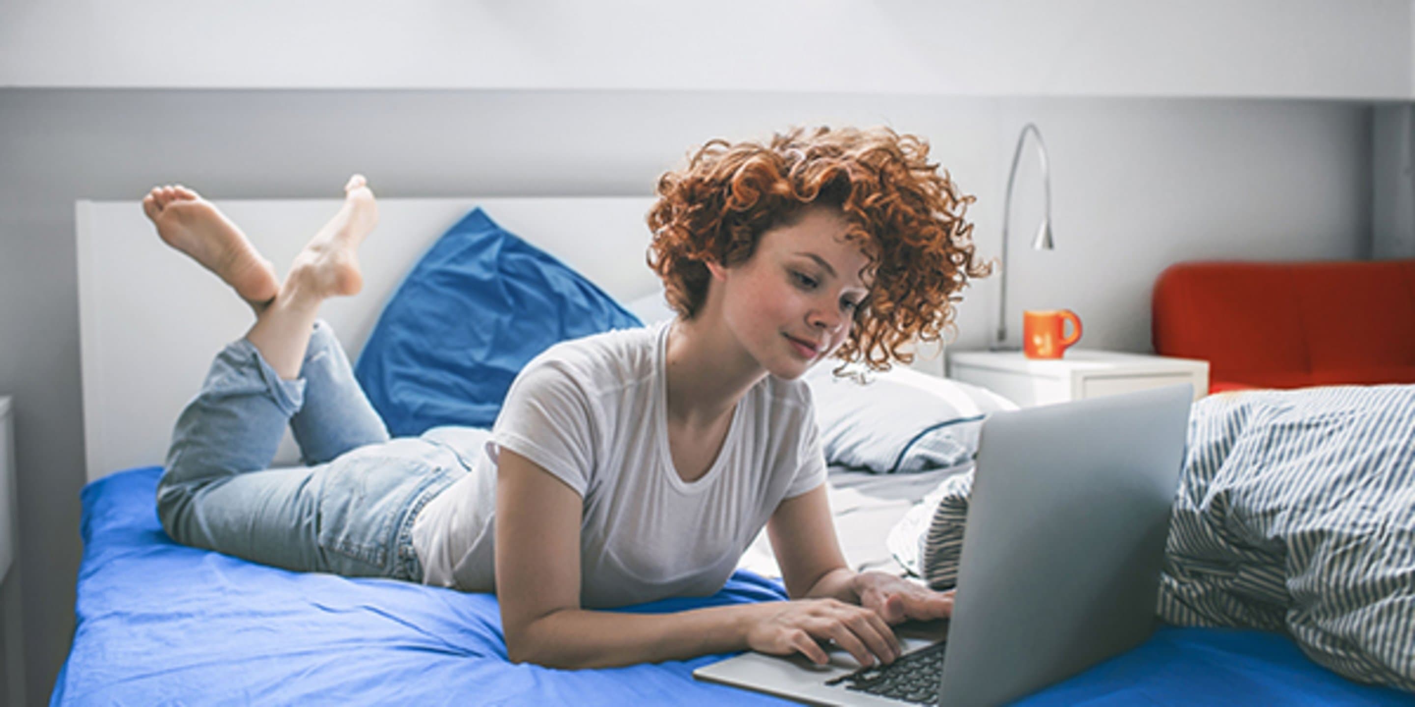 Frau mit Locken liegt auf dem Bauch falsch herum im Bett und surft am Laptop im Internet.