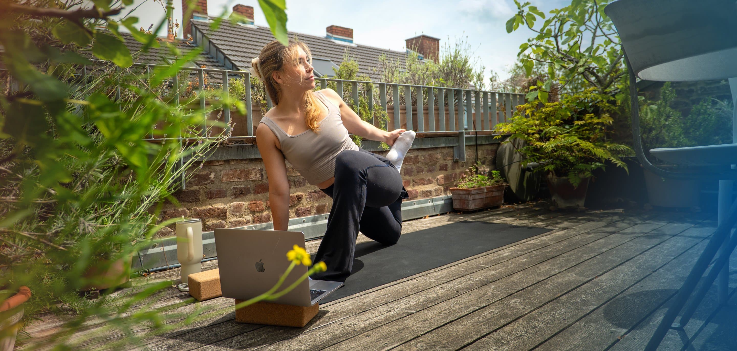 Eine Person macht Yoga auf einer schwarzen Matte auf einem Holzdeck, vor sich ein Laptop auf einem Korkblock, umgeben von Pflanzen.