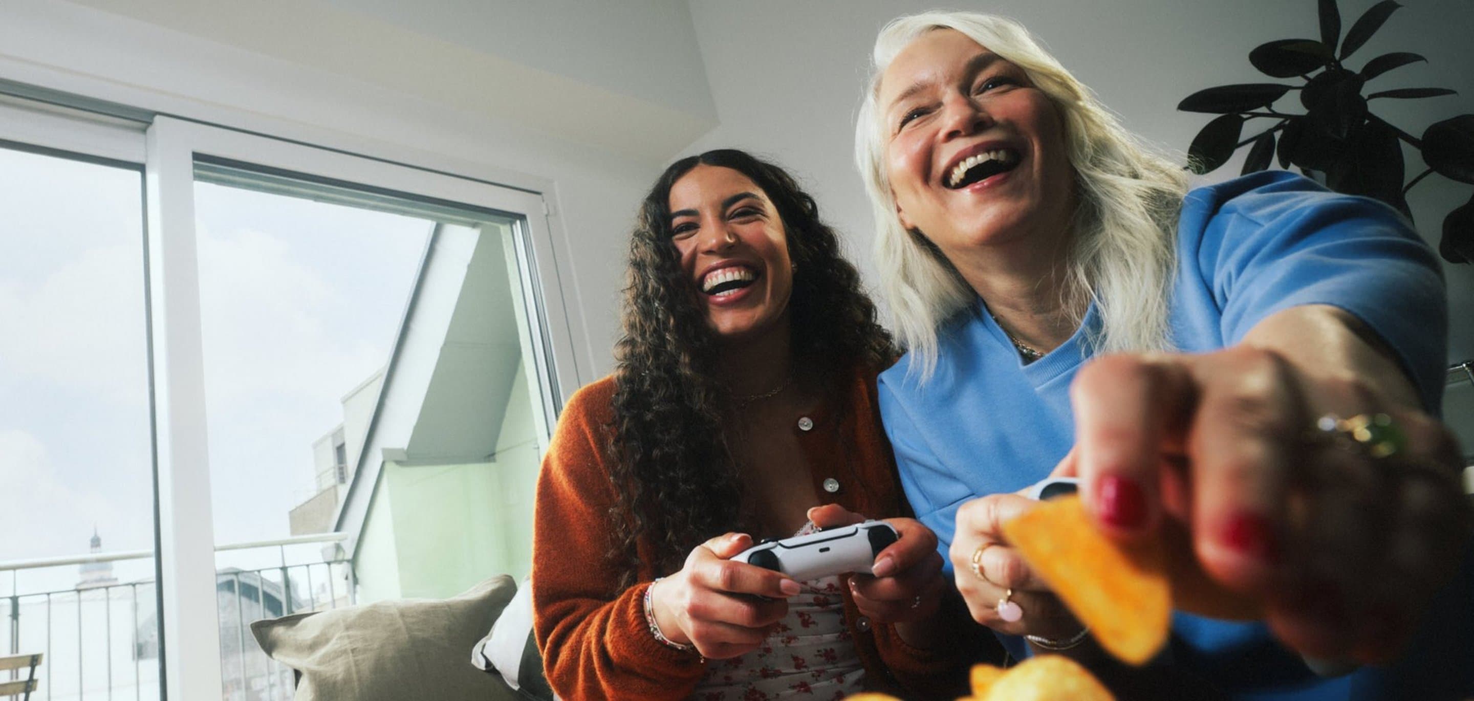 Zwei Personen sitzen in einem Wohnzimmer vor einer großen Fensterfront mit Kölnblick; eine Person hält einen Gaming‑Controller, eine andere greift nach Snacks.