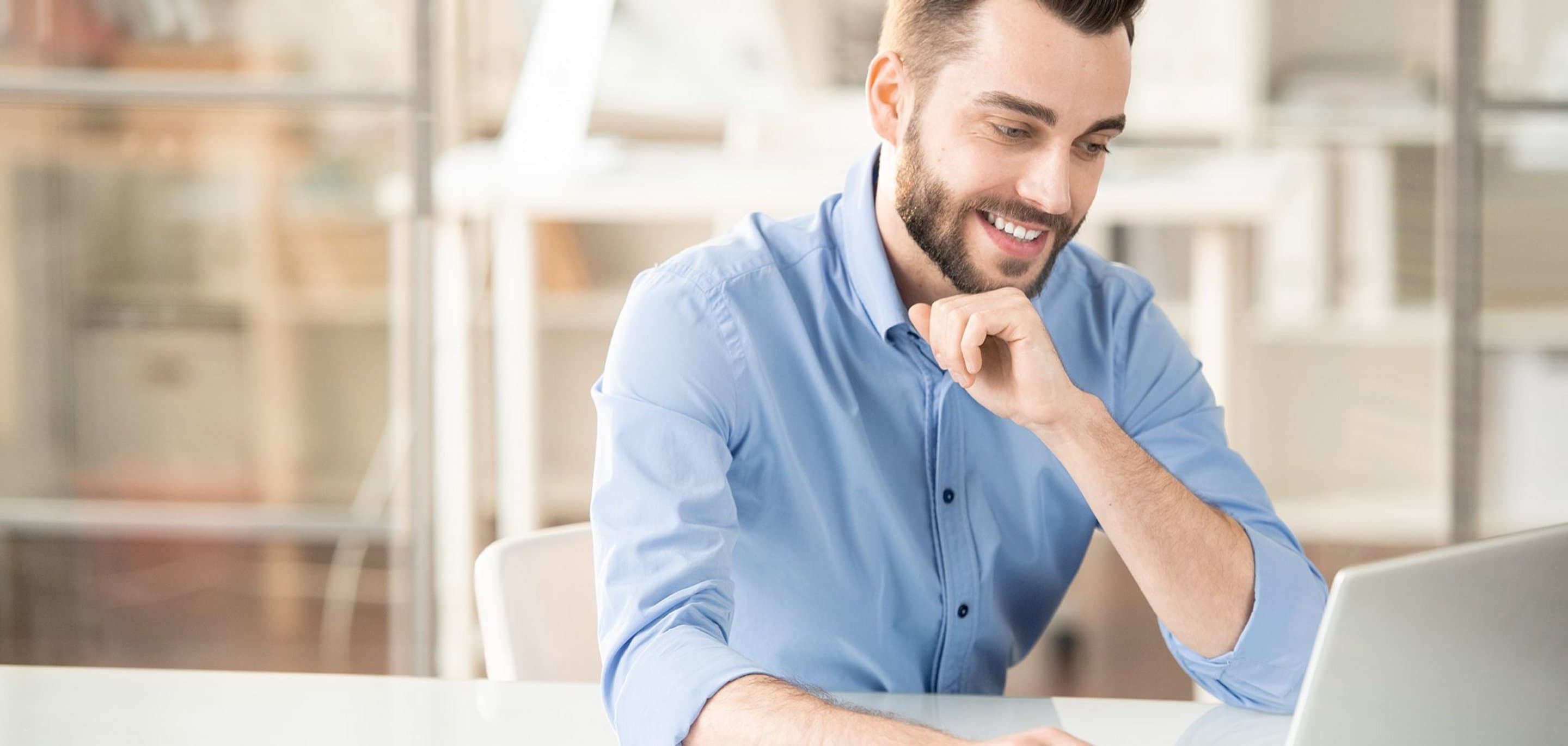 Person in hellblauem Hemd sitzt an einem Tisch vor einem Laptop in einem modernen Büro mit Glaswänden.