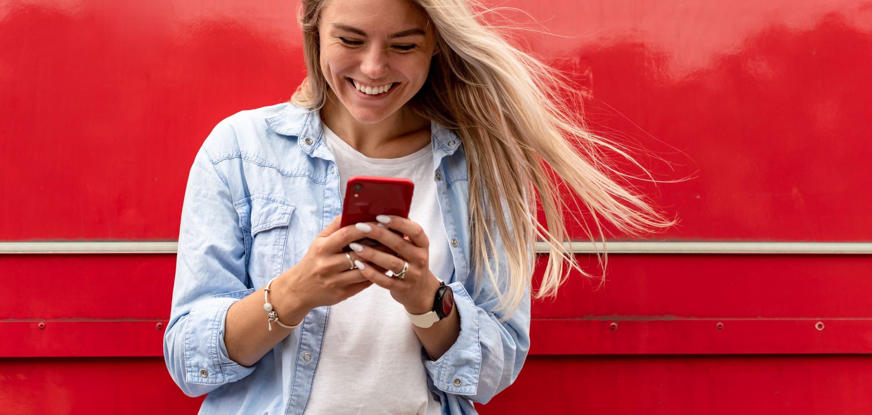 Person mit langem blondem Haar steht vor einer glänzenden roten Oberfläche und hält lächelnd ein rotes Smartphone in den Händen.