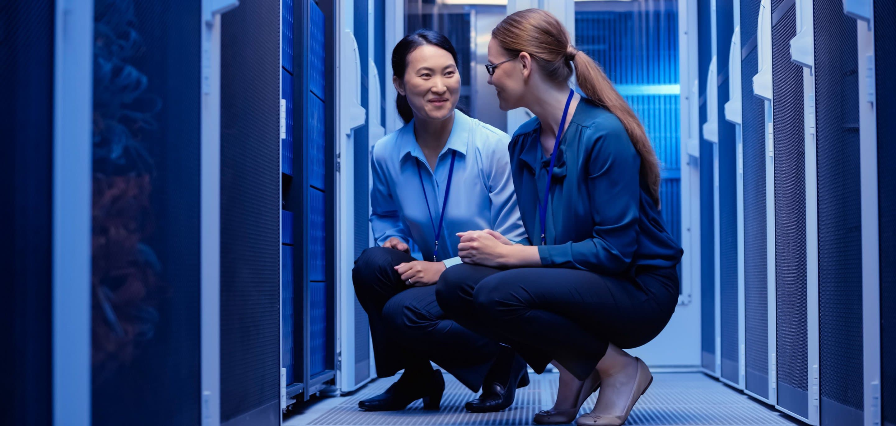 Zwei Personen in Businesskleidung knien in einem blau beleuchteten Serverraum und unterhalten sich zwischen Serverracks.
