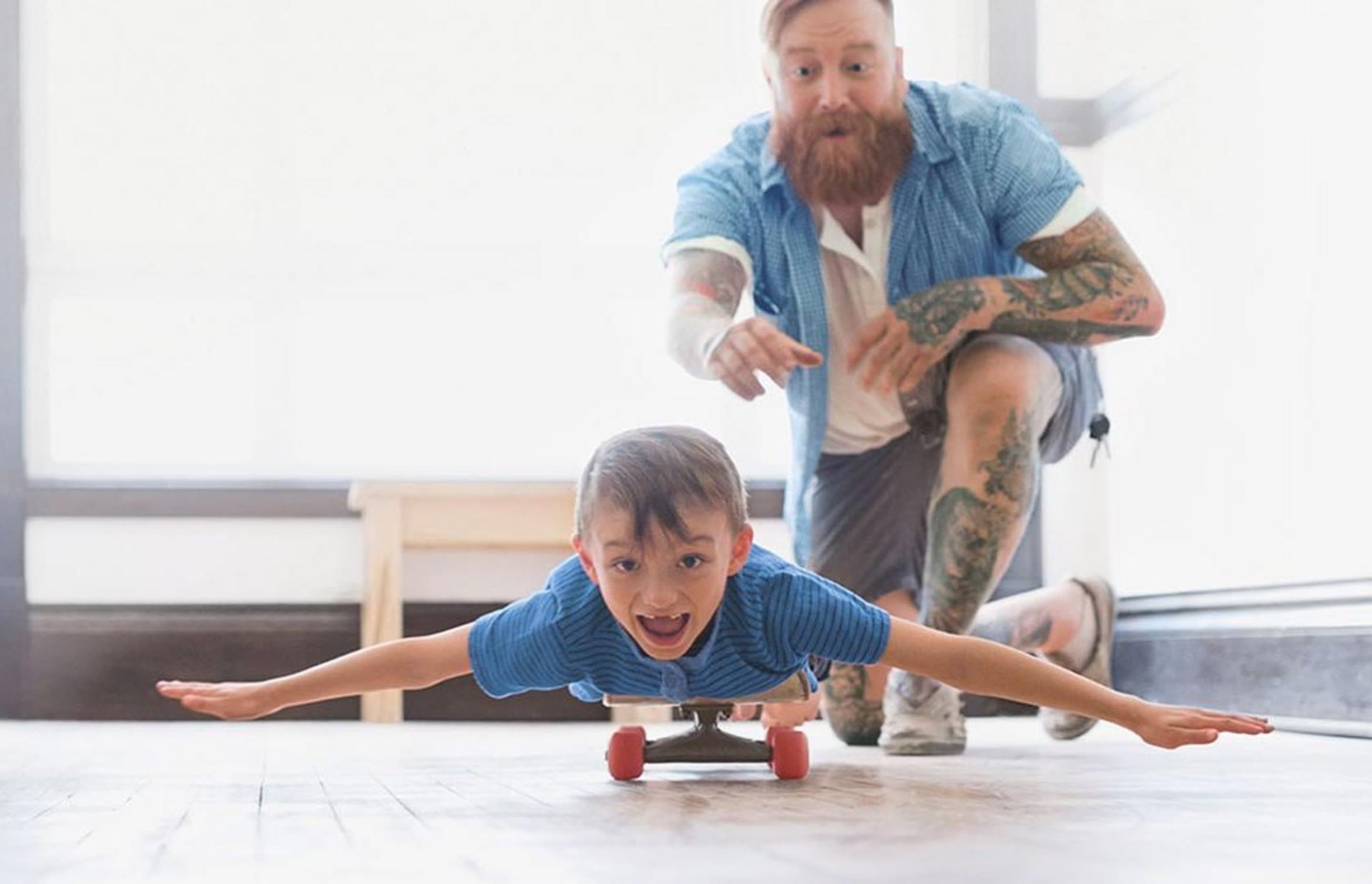 Vater schubst Sohn an, der mit ausgebreiteten Armen auf einem Skateboard liegt.