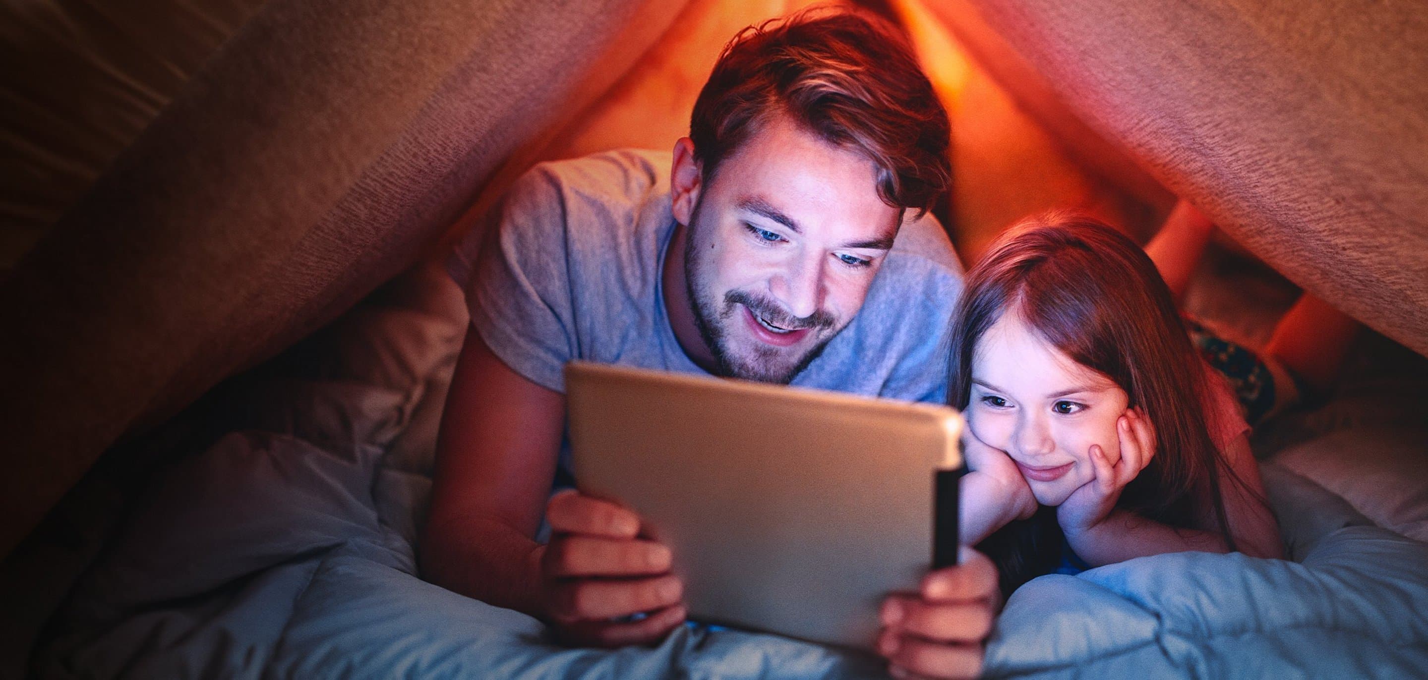 Ein Vater und seine Tochter liegen gemeinsam unter einer Decke und schauen auf ein Tablet.