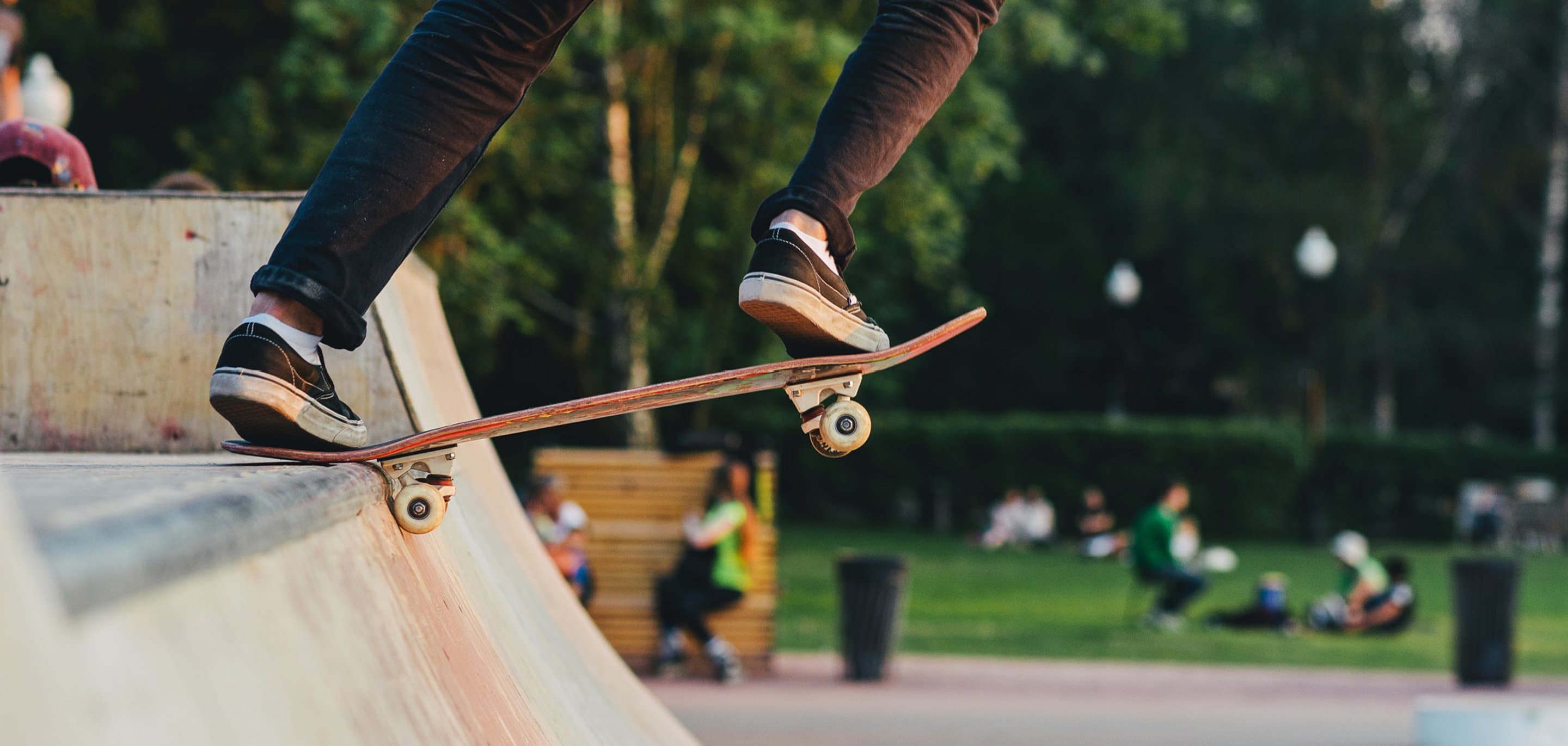 Nahaufnahme von Beinen auf einem Skateboard am Rande einer Halfpipe, im Hintergrund sind unscharf weitere Personen, Wiese und Bäume zu sehen - Symbolbild für den Skatepark North Brigade in Köln Weidenpesch.