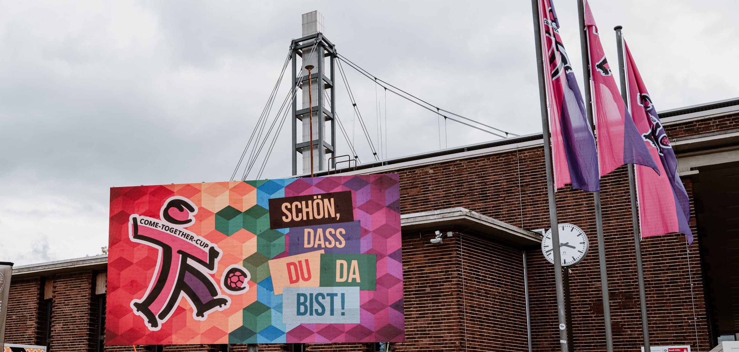 "Schön, dass du da bist." Schild des Come-Together-Cups mit dem RheinEnergieSTADION im Hintergrund.