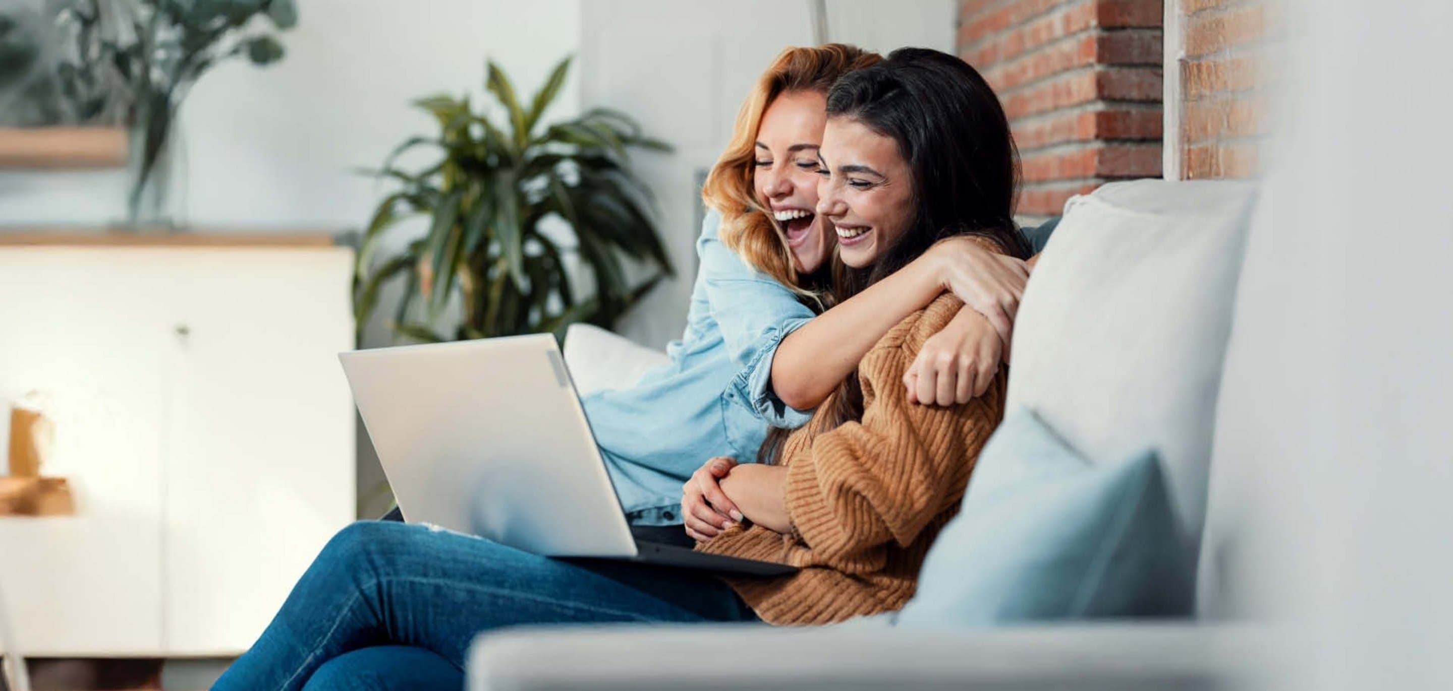 Freunde werben 200 € Praemie Zwei lachende Frauen umarmen sich auf dem Sofa und schauen gemeinsam auf einen Laptop.