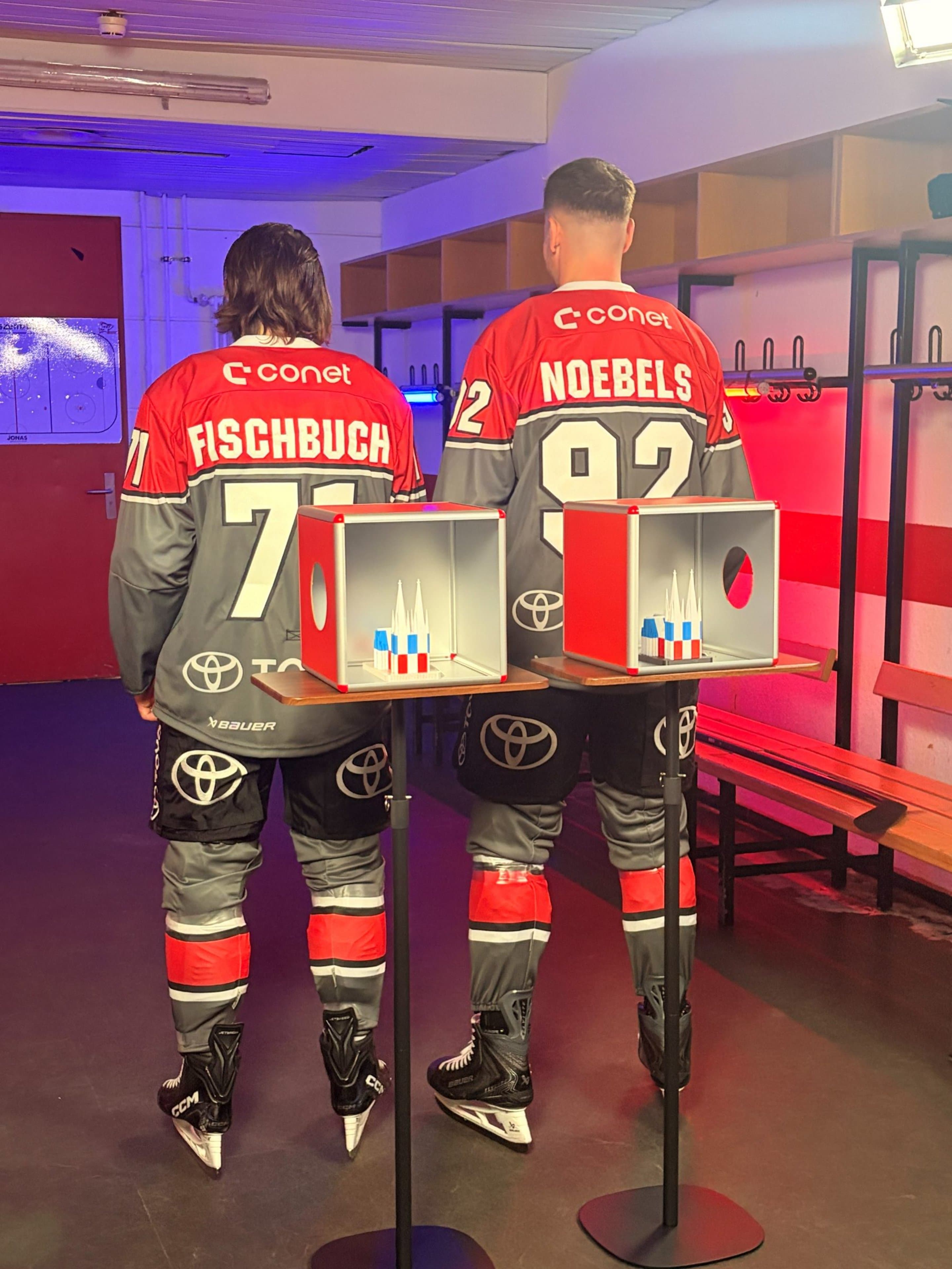 Die Kölner-Haie-Spieler Fischbuch und Noebels stehen mit dem Rücken zur Kamera. Im Vordergrund sind Miniaturen des Kölner Doms in NetCologne-Farben rot und blau zu sehen - Symbolbild für die Sponsoring-Partnerschaft zwischen Kölner Haien und NetCologne
