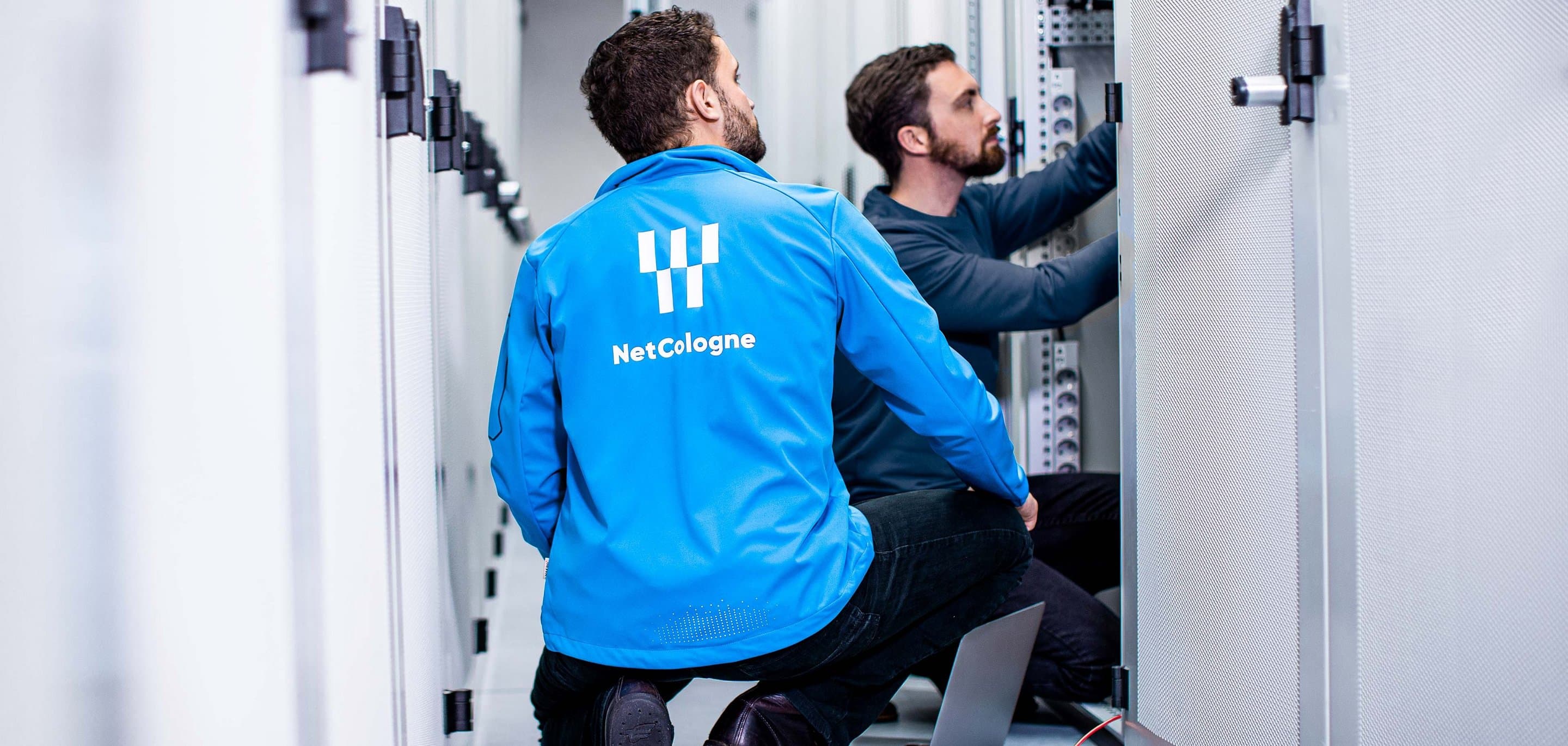 Rechenzentrum Zwei Techniker im Serverraum, einer mit „NetCologne“-Jacke, Laptop per Kabel mit Server verbunden.