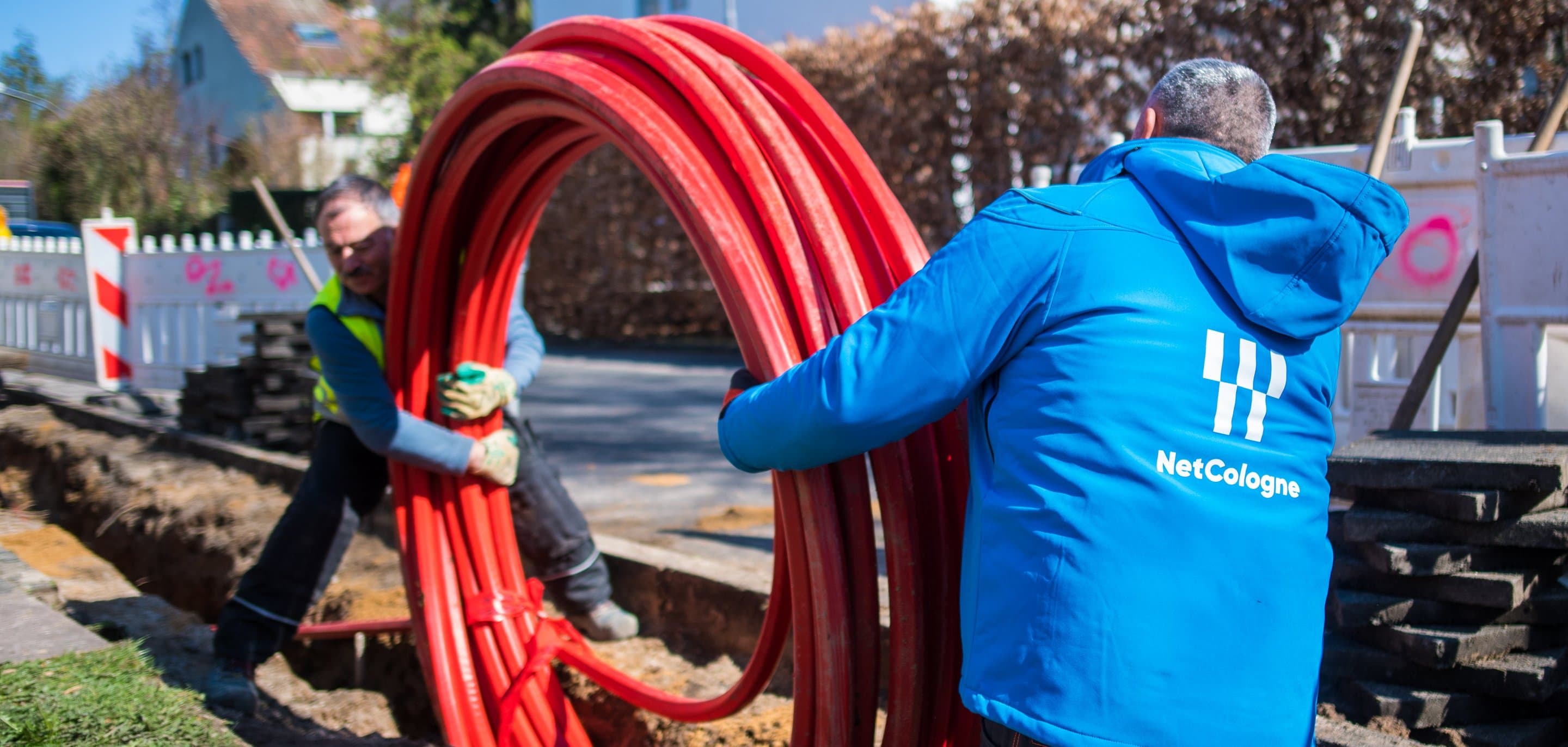 NetCologne-Mitarbeiter in blauer Jacke halten aufgerollte rote Glasfaserkabel-Leerrohre an einer Straßenbaustelle.