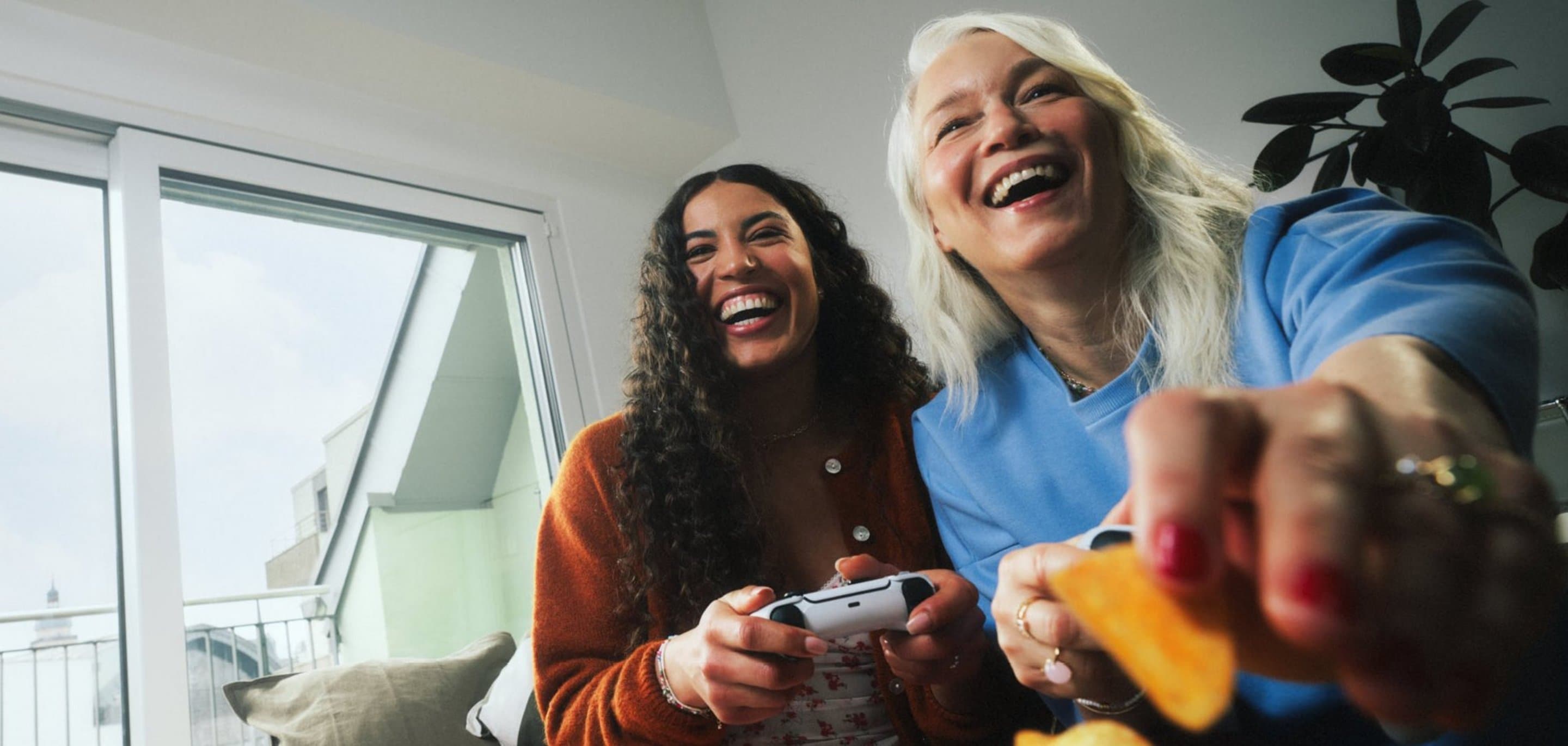 Zwei Personen sitzen in einem Wohnzimmer vor einer großen Fensterfront mit Kölnblick; eine Person hält einen Gaming‑Controller, eine andere greift nach Snacks.