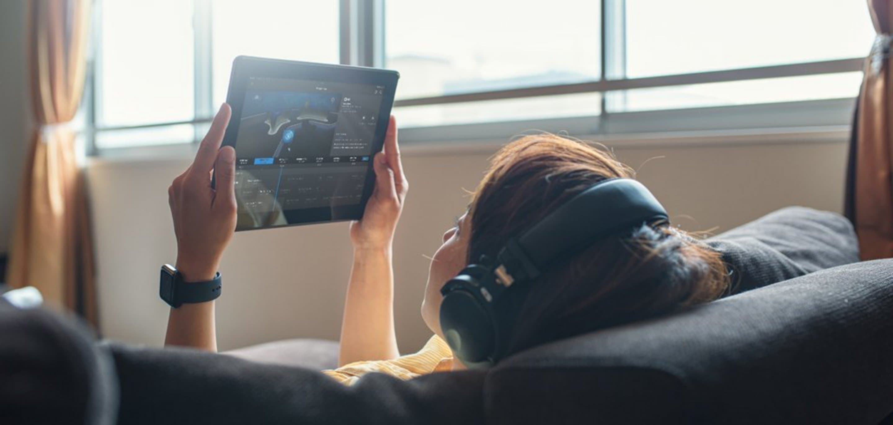 Couch von hinten auf der eine Frau liegt, die Kopfhörer trägt und Tablet in der Hand hält.