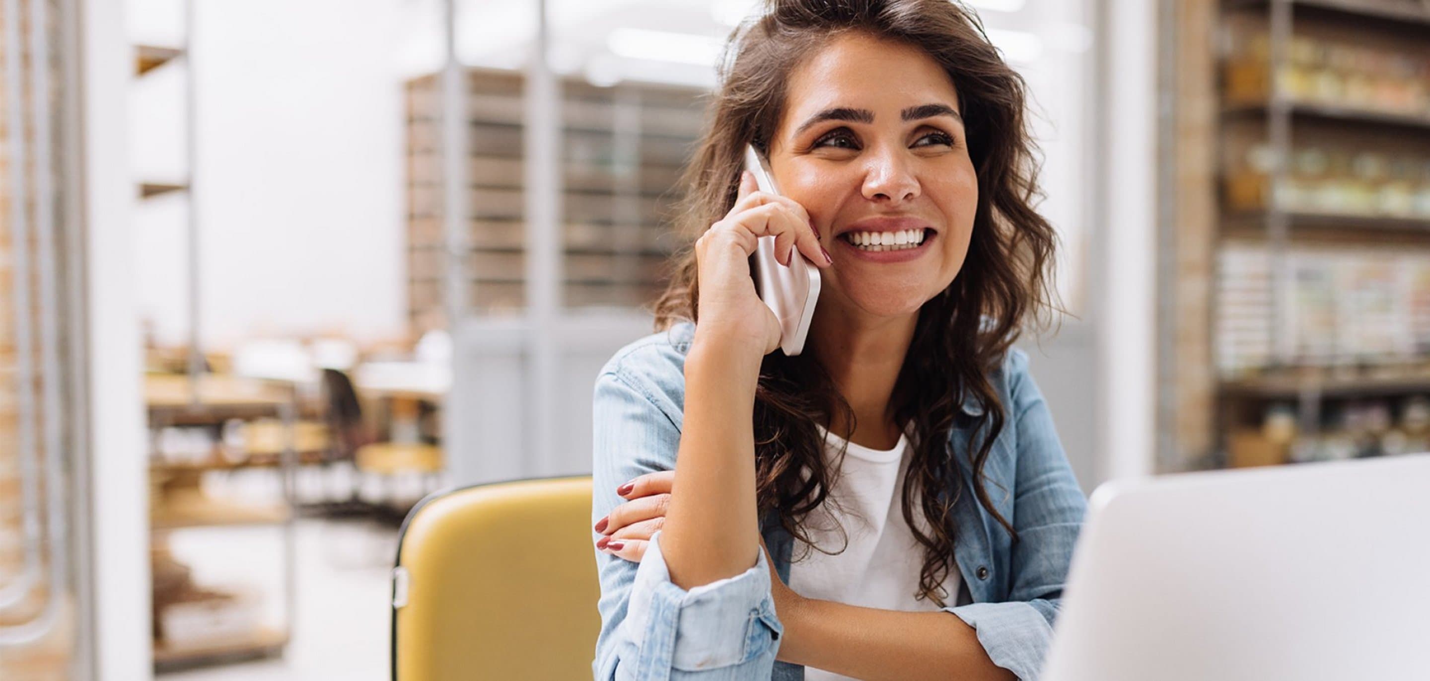 Person sitzt an einem Tisch mit Laptop und führt ein Telefonat, im Hintergrund Regale mit Büchern und Ordnern.