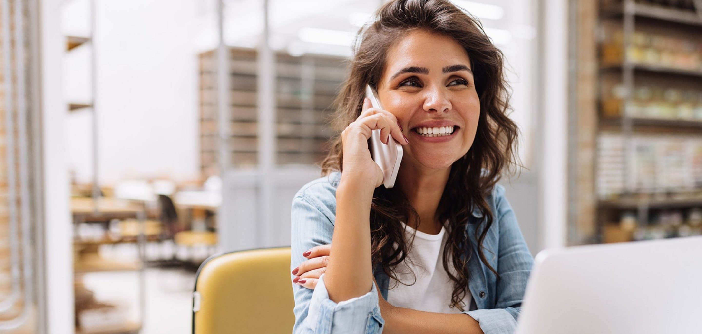 Person sitzt an einem Tisch mit Laptop und führt ein Telefonat, im Hintergrund Regale mit Büchern und Ordnern.