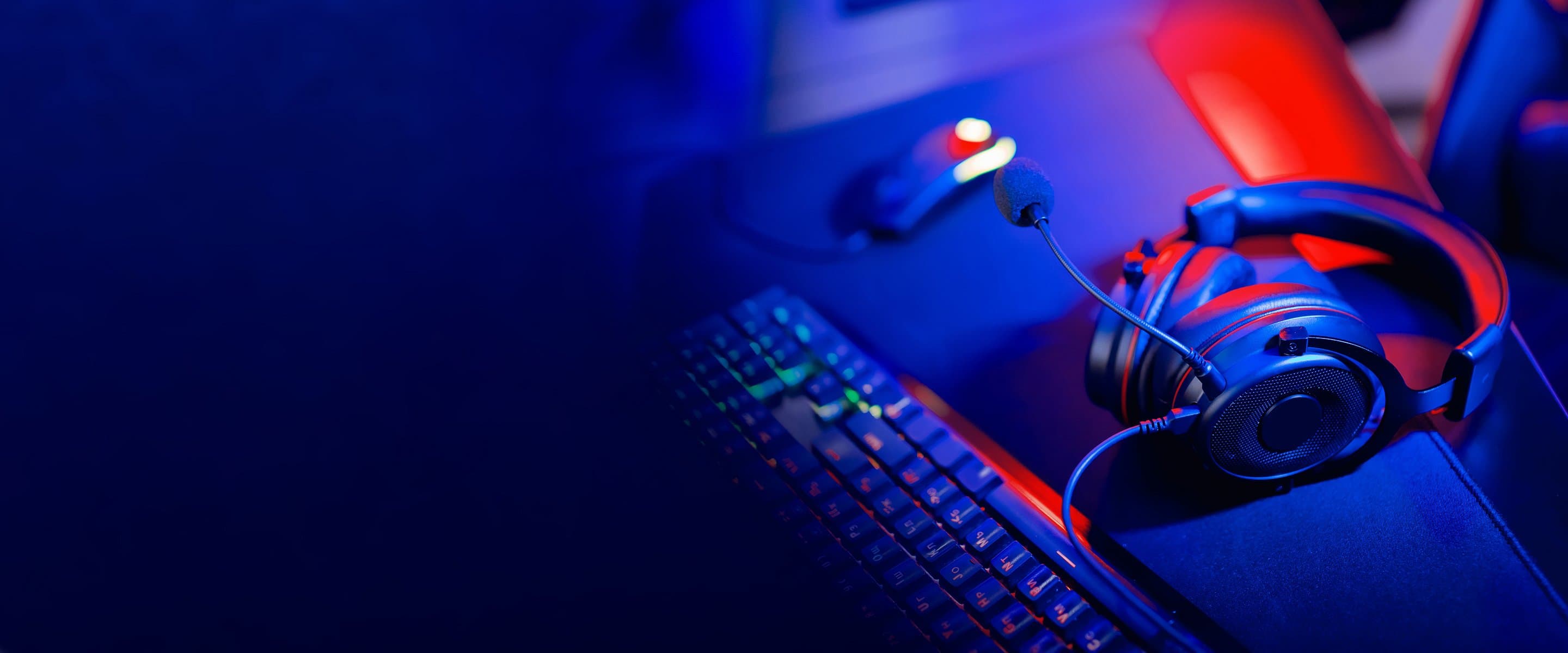 Gaming-Setup mit beleuchteter Tastatur, Computermaus und Headset mit Mikrofon, in blau-rotem Licht inszeniert für eine moderne Atmosphäre.