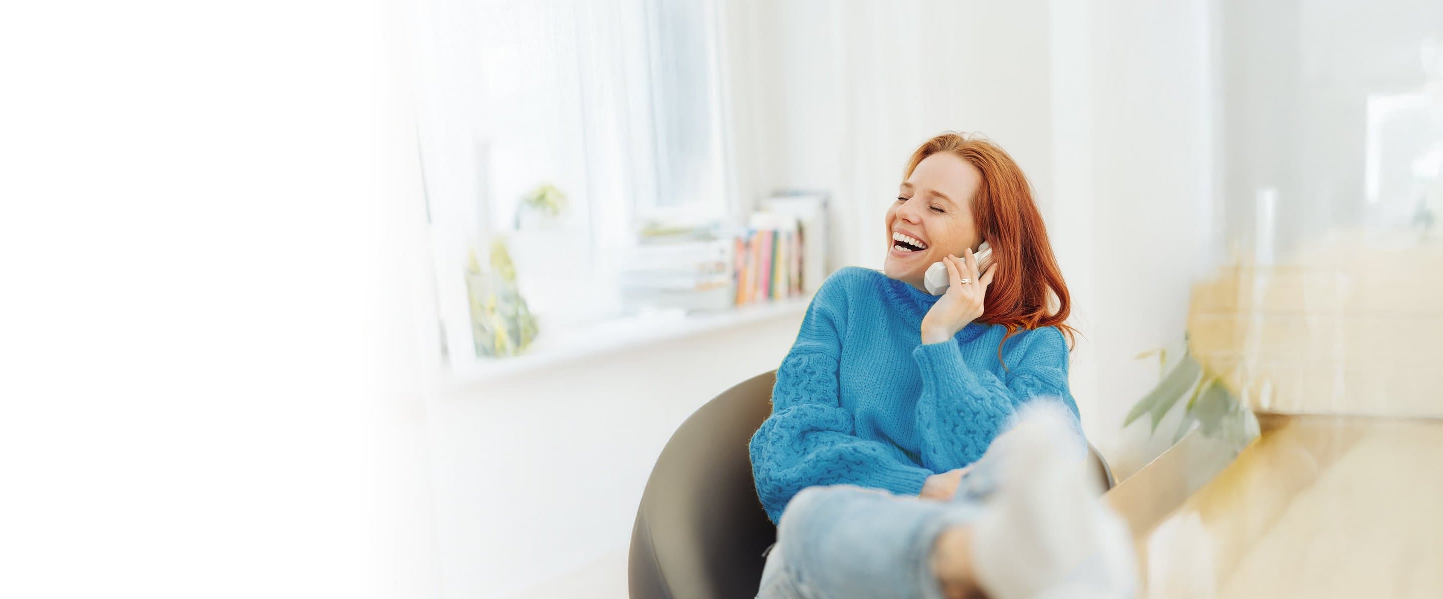 Lächelnde Frau mit rotem Haar sitzt entspannt in einem Sessel und telefoniert über ein Festnetztelefon in einem hellen, gemütlichen Raum.