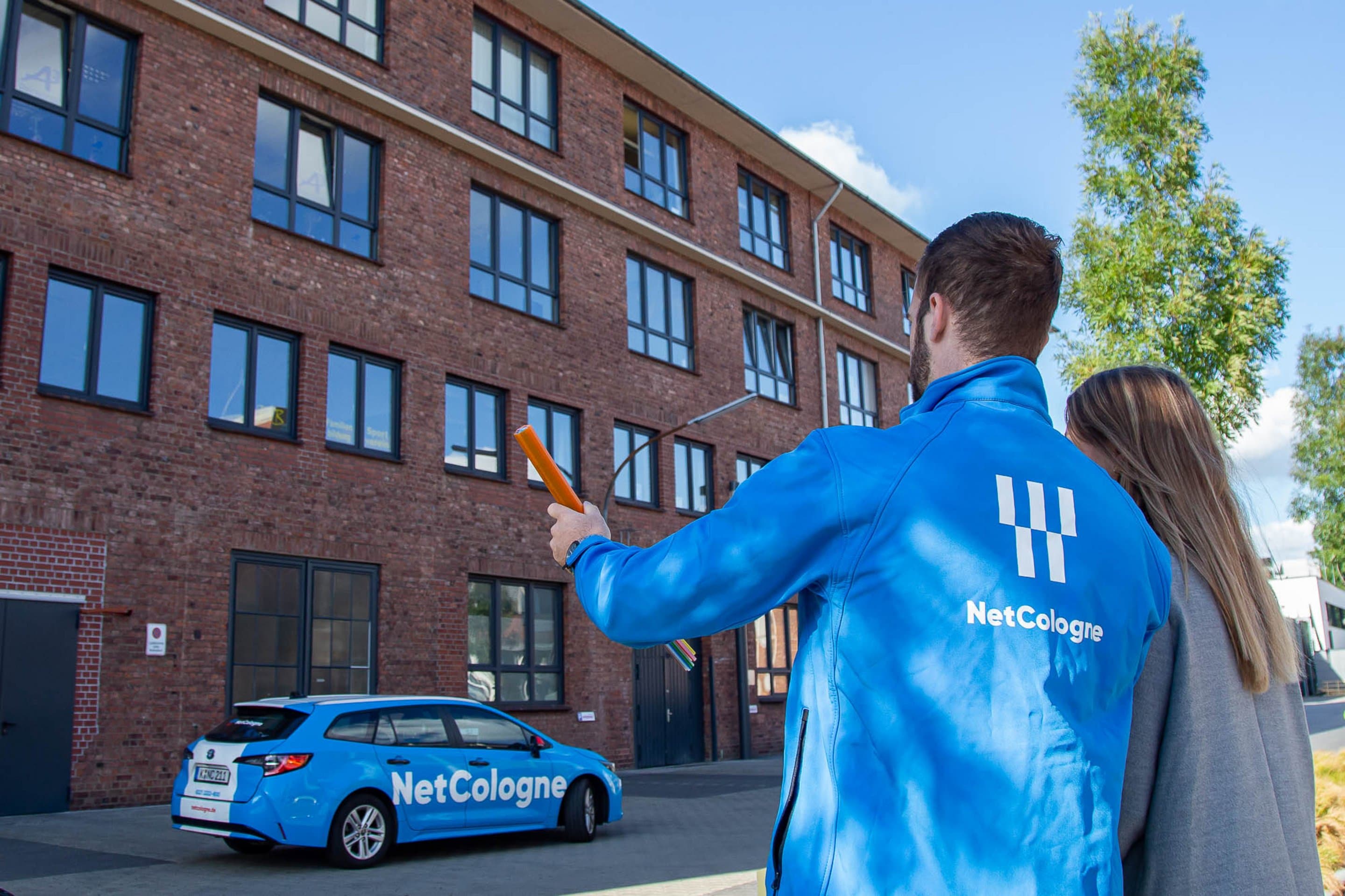 Symbolbild für Landingpage Glasfaser-Tarife Zwei Personen stehen vor einem Backsteingebäude, vor dem ein blaues Auto mit NetCologne-Schriftzug parkt.