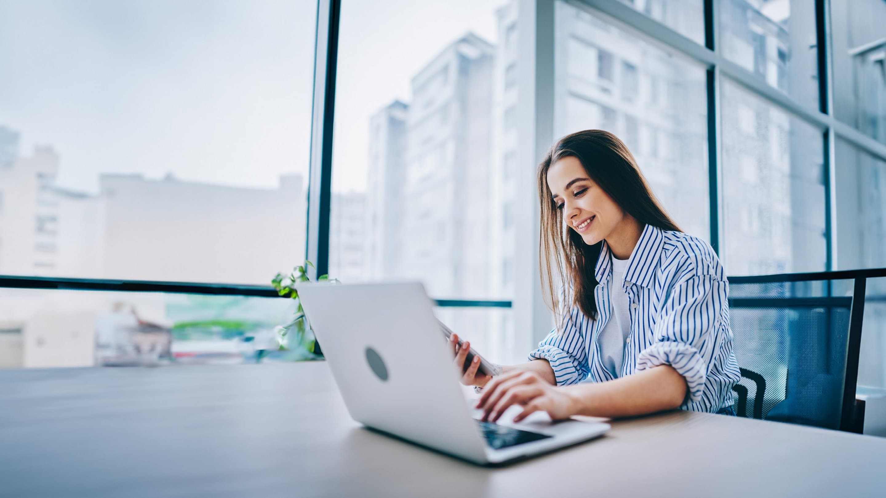 Sichere & werbefreie E-Mail erstellen mit NetMail Frau sitzt am Schreibtisch auf dem ein Laptop steht und schaut auf ihr Handy