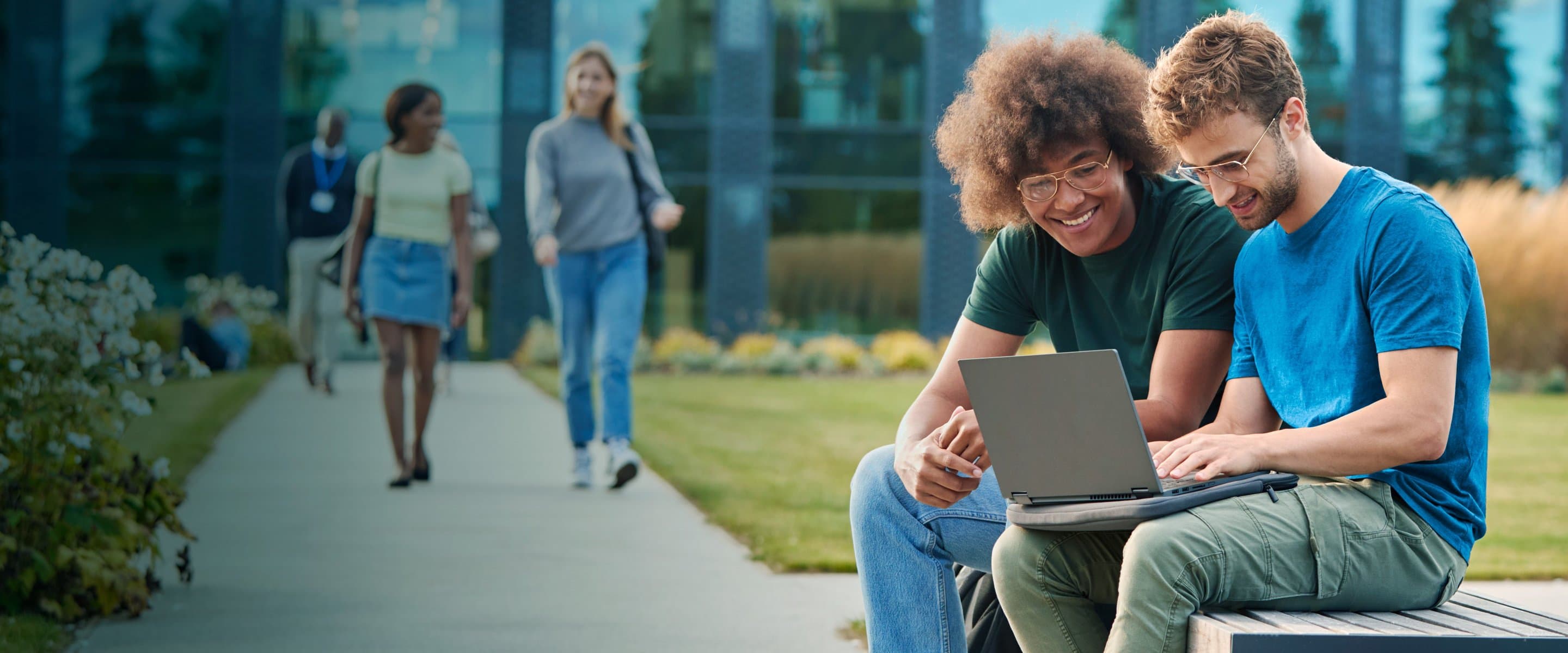 Zwei junge Männer sitzen auf einer Bank im Freien und schauen gemeinsam lächelnd auf einen Laptop, während im Hintergrund Menschen auf einem Weg spazieren.