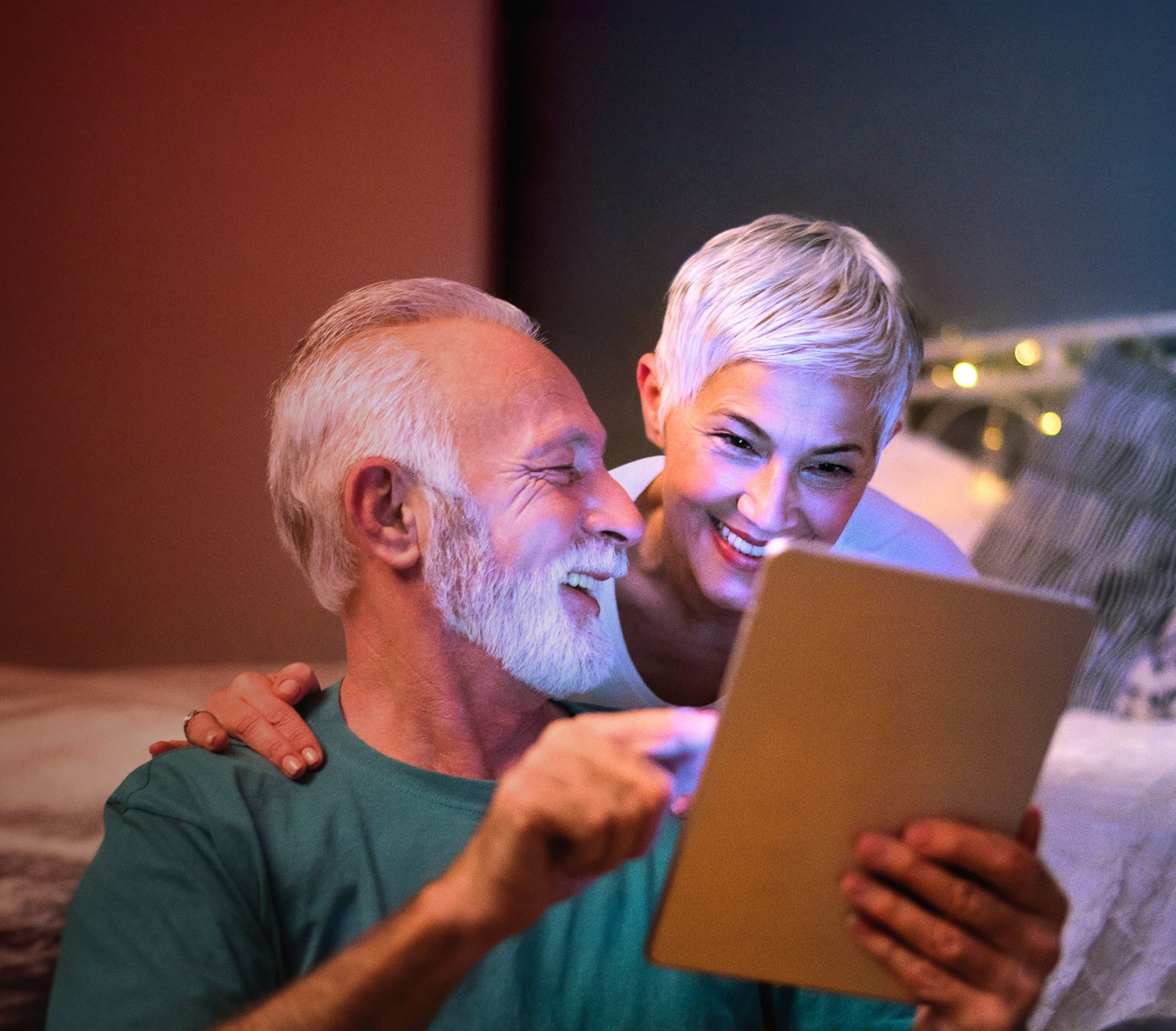 Zwei ältere Personen sitzen nebeneinander. Eine Person hält ein Tablet in den Händen. Im Hintergrund ist ein Bett mit Kissen und Lichterkette zu sehen.