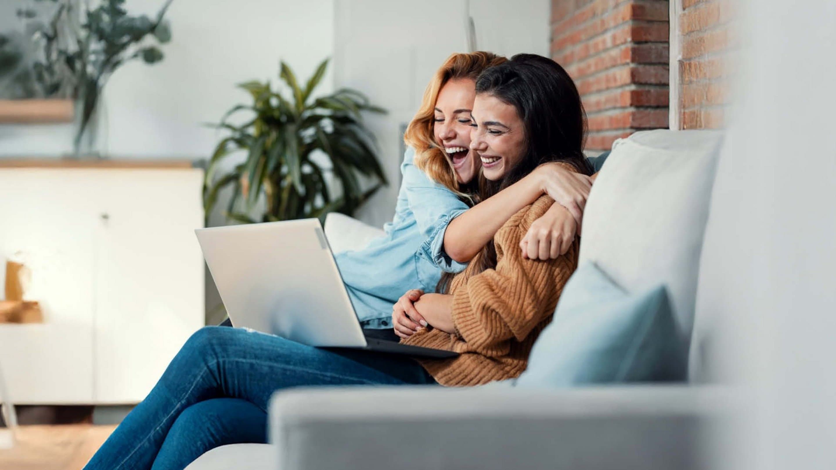 Freunde werben 200 Euro Praemie Zwei lachende Frauen umarmen sich auf dem Sofa und schauen gemeinsam auf einen Laptop.