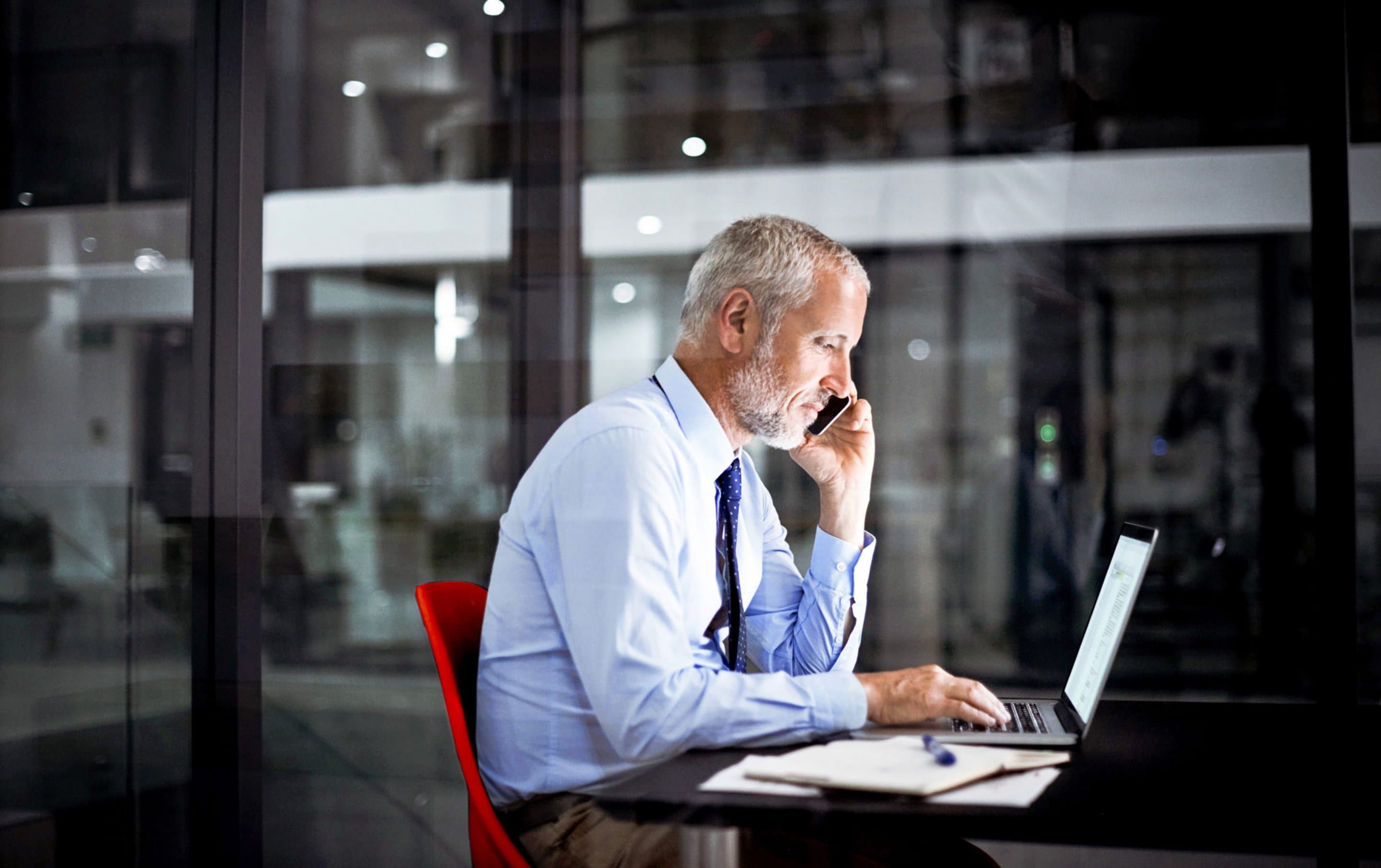 Business Internet Person in hellblauem Hemd sitzt an einem schwarzen Tisch vor einem Laptop, daneben ein Notizbuch und ein Stift, im Hintergrund Glaswände eines modernen Büros.